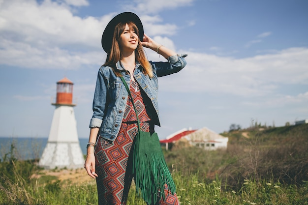 Free photo young woman in nature, lighthouse, bohemian outfit, denim jacket, black hat, smiling, happy, summer, stylish accessories