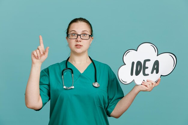 Young woman nurse in medical uniform with stethoscope around neck holding bubble speech with word idea looking confident showing index finger standing over blue background