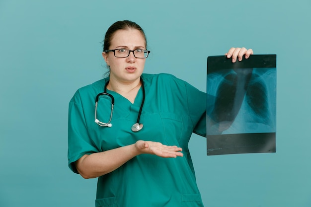 Young woman nurse in medical uniform with stethoscope around neck holding lung xray presenting with arm of her hand looking confused having doubts standing over blue background