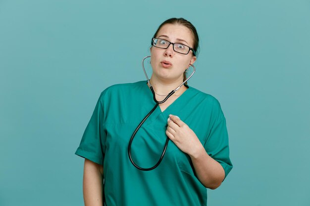 Young woman nurse in medical uniform with stethoscope around neck looking at camera amazed and surprised standing over blue background