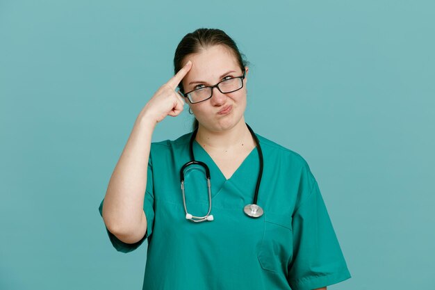 Young woman nurse in medical uniform with stethoscope around neck looking at camera confused pointing with index finger at her temple for mistake standing over blue background