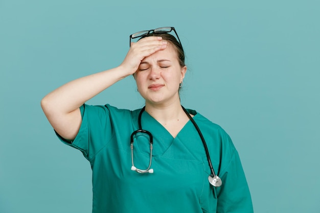 Young woman nurse in medical uniform with stethoscope around neck looking tired and overworked holding hand on her forehead standing over blue background