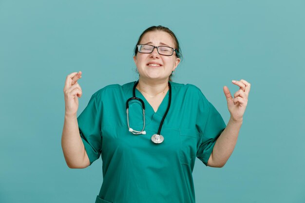 Young woman nurse in medical uniform with stethoscope around neck making wish crossing fingers with hope expression standing over blue background