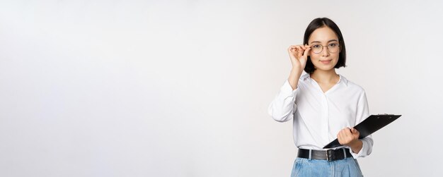 Young woman office worker manager in glasses holding clipboard and looking like professional standing against white background