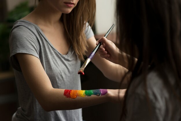 Free Photo young woman painting the rainbow flag over her girlfriend's hand with paintbrush