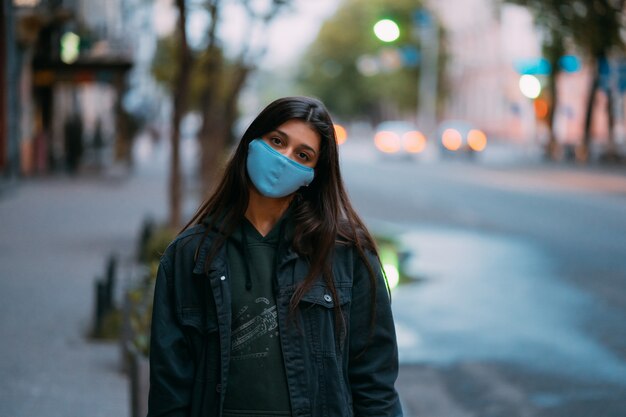 Young woman, person in protective medical sterile mask standing at empty street, looking at camera.