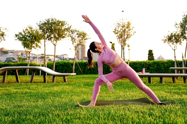 young woman in pink shirt and trousers on the grass inside green park meditating and doing yoga in different poses