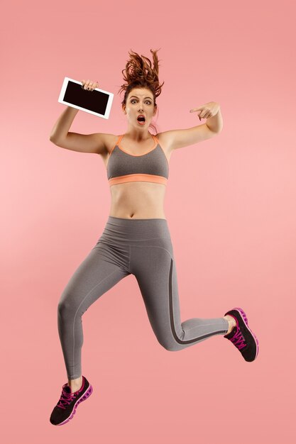 Young woman over pink studio wall using tablet gadget while jumping