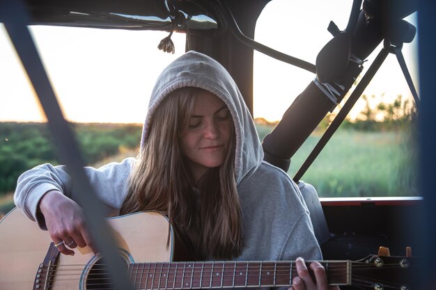 A young woman plays the acoustic guitar in the trunk of a car in nature