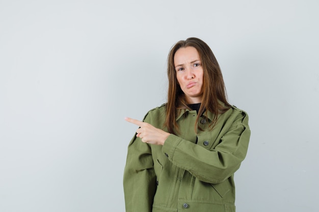 Free photo young woman pointing aside in green jacket and looking unhappy , front view.