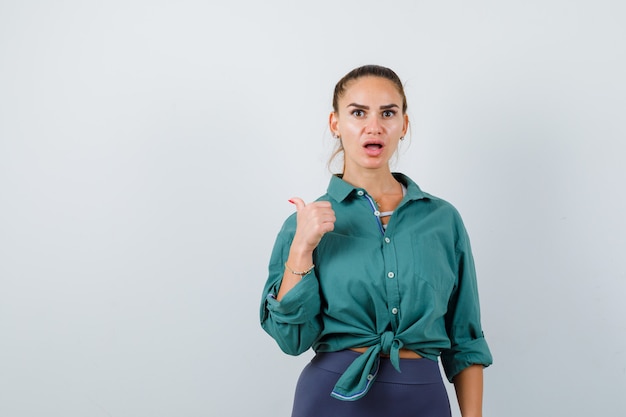 Young woman pointing back with thumb in green shirt and looking surprised , front view.