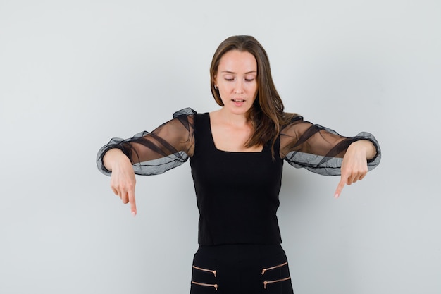 Free photo young woman pointing down with both index fingers and looking down in black blouse and black pants and looking optimistic