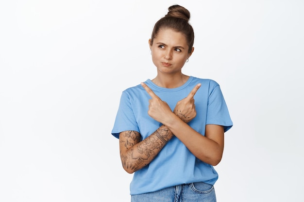 Free Photo young woman pointing fingers sideways and looking left, making her choice between two options, standing in blue tshirt over white background.