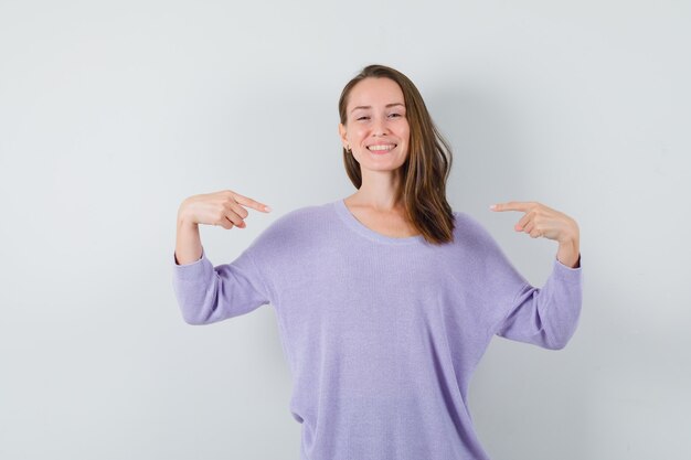 Young woman pointing at herself in lilac blouse and looking optimistic. front view.