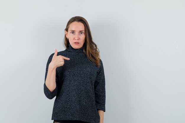 Young woman pointing at herself in shirt and looking puzzled. front view.