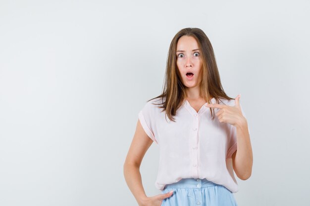 Young woman pointing at herself in t-shirt, skirt and looking shocked. front view.