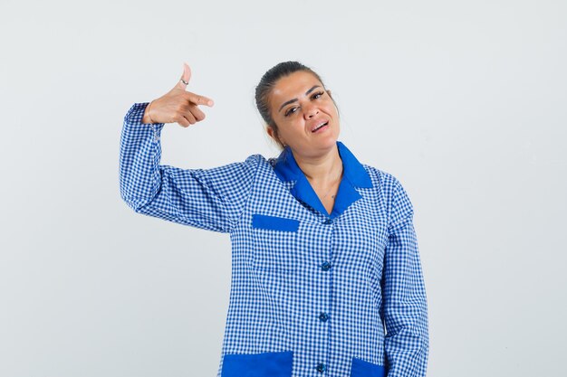 Young woman pointing at herself with index finger in blue gingham pajama shirt and looking pretty , front view.