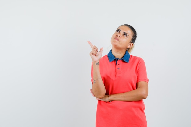 Young woman pointing left with index finger in red shirt and looking pensive. front view.