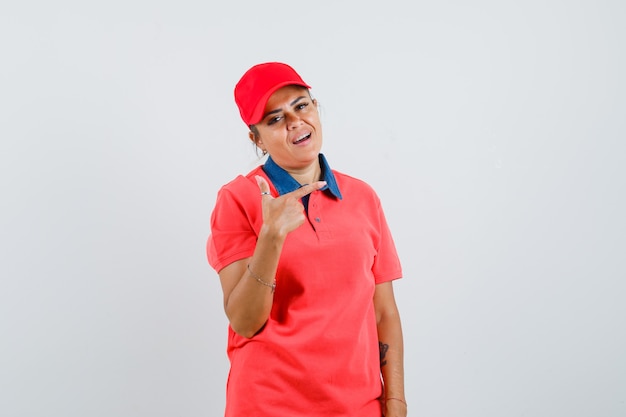 Young woman pointing right with index finger in red shirt and cap and looking pretty. front view.