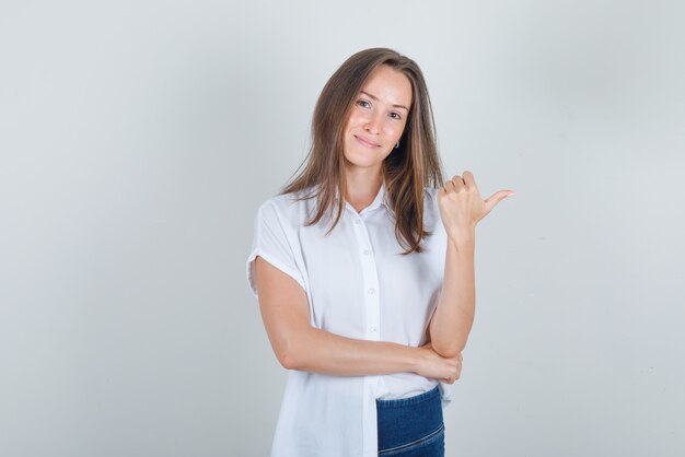 Young woman pointing to side with thumb up in t-shirt, jeans and looking cheerful