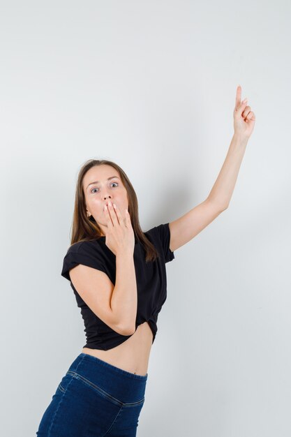 Young woman pointing up while covering her mouth with hand in black blouse and looking scared.