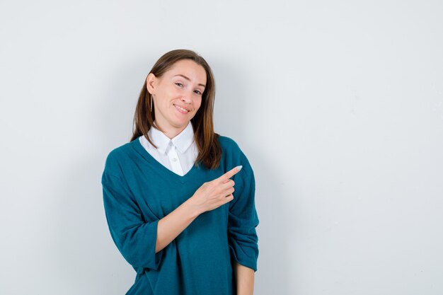 Young woman pointing at upper right corner in sweater over white shirt and looking jolly , front view.