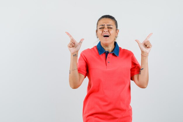 Young woman pointing upward with index fingers in red t-shirt and looking amused. front view.