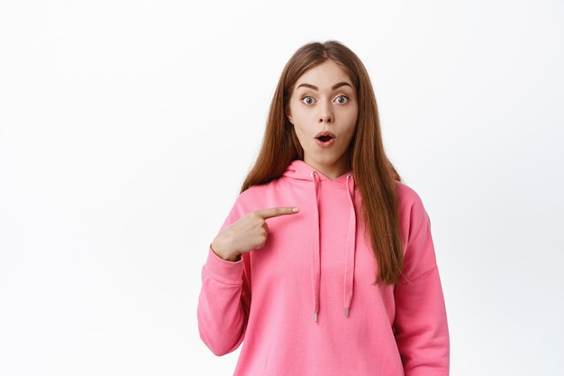 Young woman points at herself, gasps and looks surprised, being chosen, picked to participate, stare amazed at front, stands against white wall