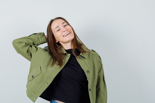 Young woman posing with hand behind head in green jacket and looking jolly. front view.