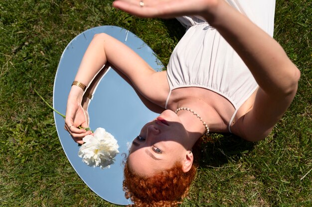 Young woman posing with peony flower in the mirror on the grass outdoors