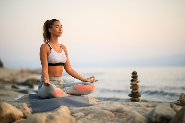 Young woman practicing Yoga relaxation exercises and meditating on a rock at the beach. Copy space.