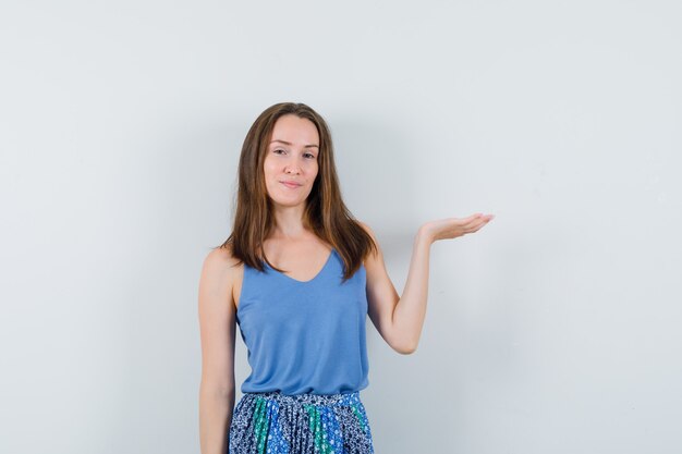 Young woman pretending to hold or show something in singlet, skirt and looking confident , front view.