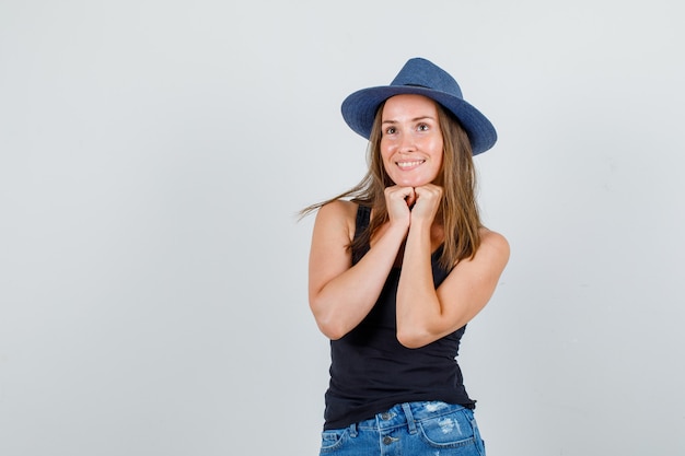 Young woman propping chin on fists in singlet, shorts, hat and looking hopeful