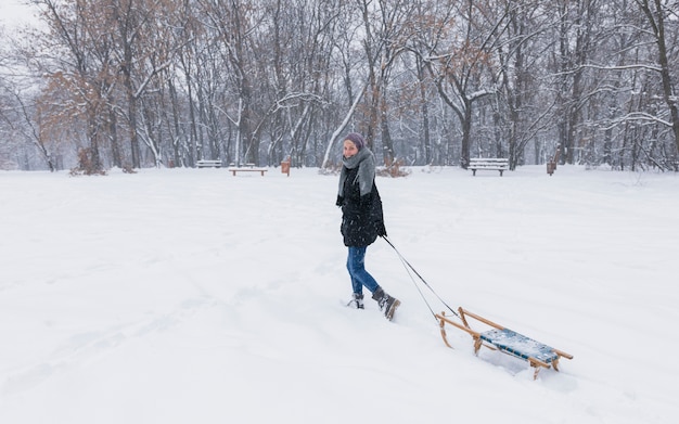 Young woman pulling empty wooden sledge on snow landscape at forest