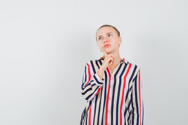 Young woman putting her fingers on chin and looking upward in striped blouse and looking doubtful