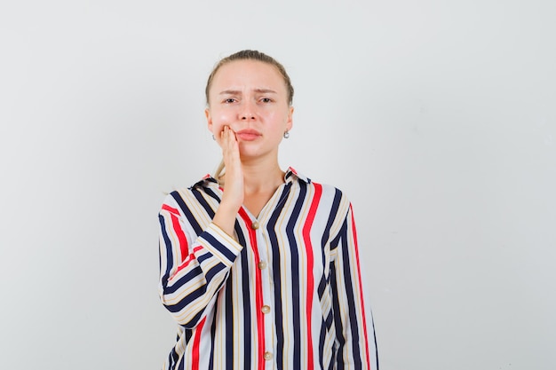 Young woman putting her hand on cheek in striped blouse and looking confused