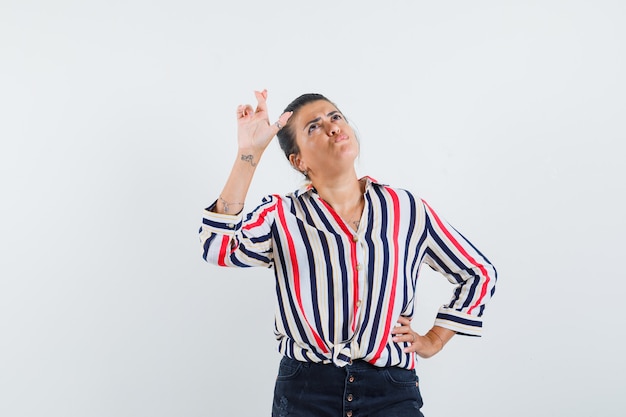 Young woman putting one hand on waist, standing finger crossed and thinking about something in striped blouse and looking pensive