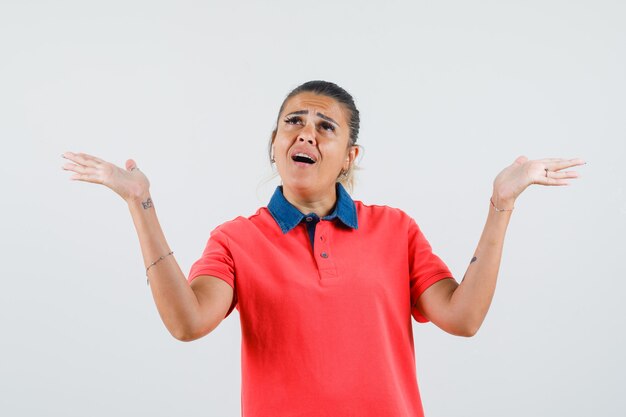 Young woman raising hands as holding something in red t-shirt and looking surprised. front view.