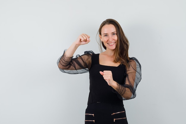 Young woman raising her fists in black blouse and looking confident. front view.