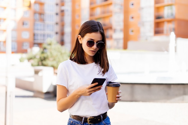 Free photo young woman reading using phone. female woman reading news or texting sms on smartphone while drinking coffee on break from work.