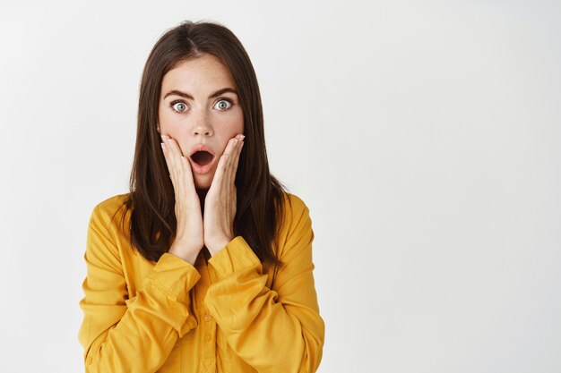 Young woman receive shocking bad news, staring at camera startled, touching face and drop jaw, standing over white wall.