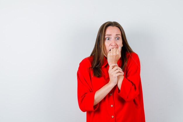 Young woman in red blouse with fist on mouth and looking scared , front view.