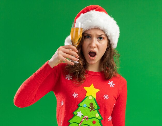 young woman in red christmas sweater and santa hat holding glass of champagne looking at camera annoyed and irritated standing over green background