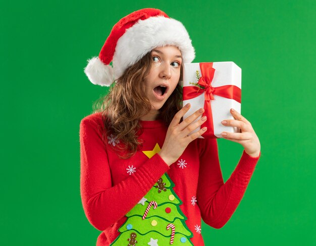 Young woman in red christmas sweater  and santa hat holding  a present  being amazed and surprised standing over green  background