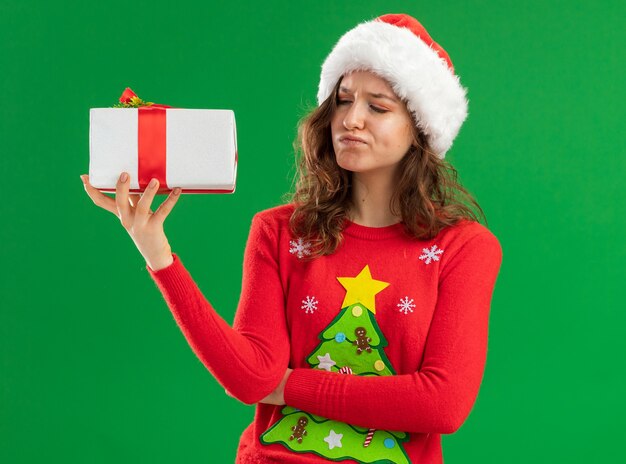 Young woman in red christmas sweater  and santa hat holding  a present looking at it with skeptic expression  standing over green  background