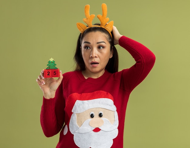 Free Photo young woman in  red christmas sweater wearing funny rim with deer horns   holding toy cubes with date twenty five looking aside confused and worried  standing over green wall