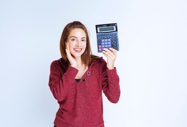 Young woman in red jacket calculating something on a blue calculator and demonstrating the final result