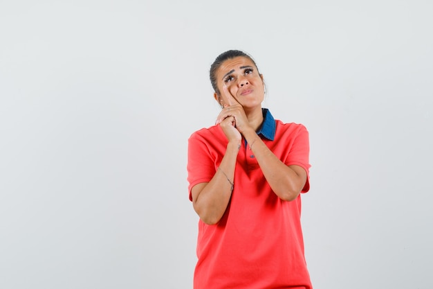 Young woman in red t-shirt leaning cheek on hand and looking pensive , front view.