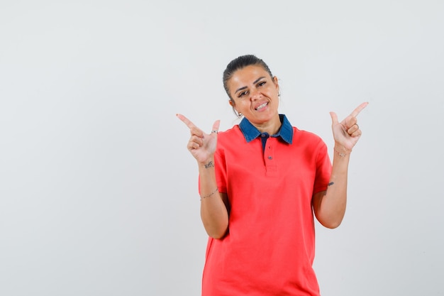 Young woman in red t-shirt pointing at different directions with index fingers and looking pretty , front view.