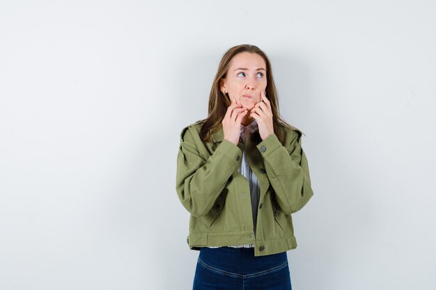 Young woman in shirt, jacket holding hands on chin and looking pensive , front view.